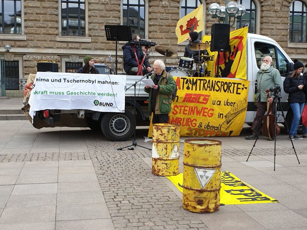Redner von SAND am Rathausmark, mit gelben Atomfässern im Vordergrund und Wagen mit Musik und weiteren Antiatom Banner im hintergrund