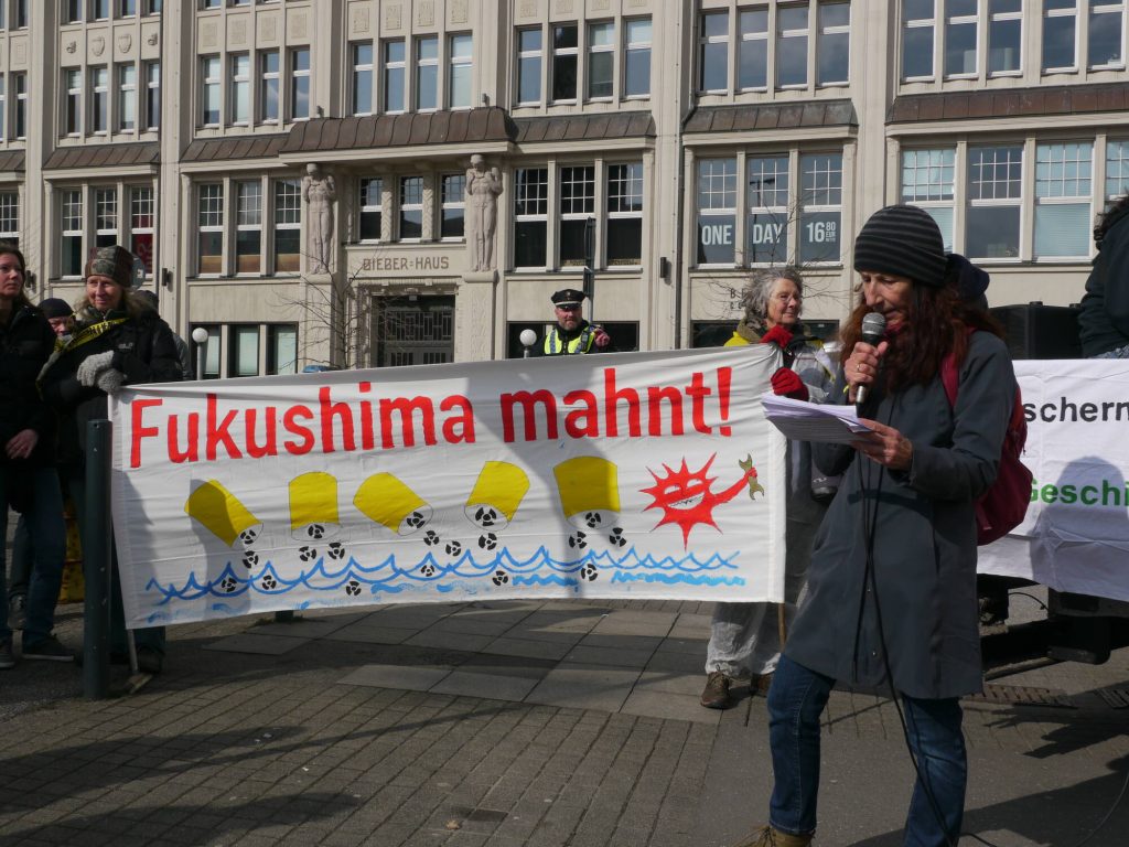 eine Person spricht ins Mikrofon mit Banner Fukushima Mahnt im Hintergrund