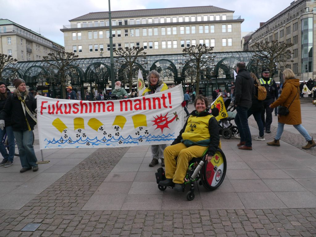 Banner Fukushima Mahnt und Demonstrierende, eine Person mit Rollstuhl und antiatom Fahne sitzt rechs vom Banner